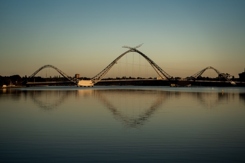horizontal shot of a bridge with wavy structures reflecting in a body of water with clear skies
