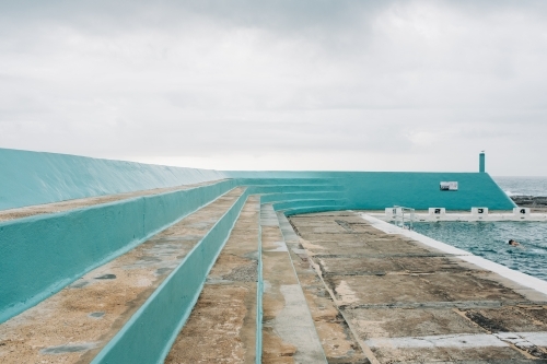 horizontal shot of a concrete ocean pool bleacher with a person swimming on a cloudy day