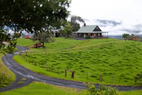 horizontal shot of a house with green lawn and foggy mountains in the background and trees in front - Australian Stock Image