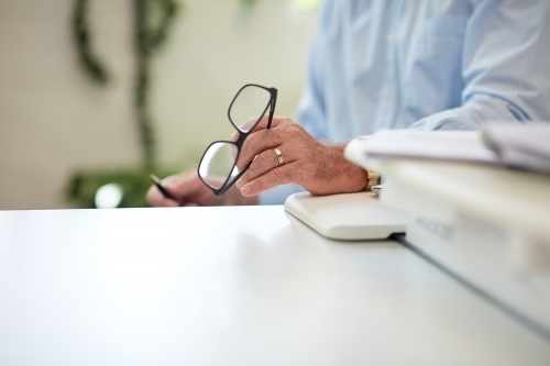 Horizontal shot of a man's left hand resting on a white desk holding his reading glasses - Australian Stock Image