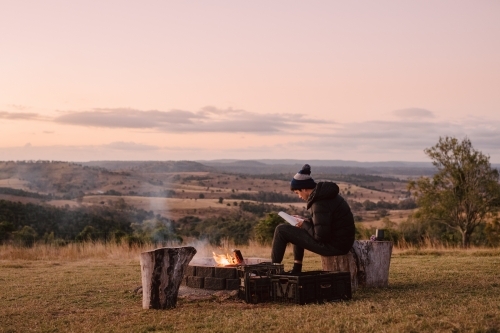 Horizontal shot of a man sitting on a log near a camp fire on a hill top while reading a book