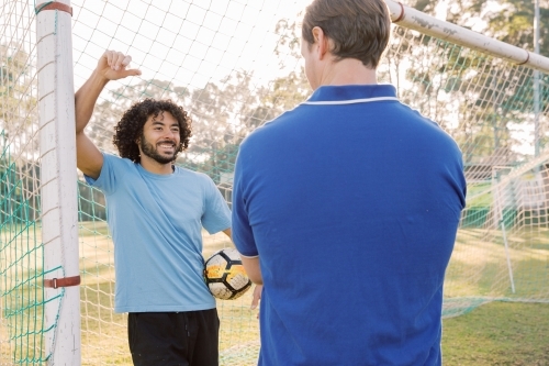 horizontal shot of a man with a soccer ball smiling to another man while standing against a goal - Australian Stock Image