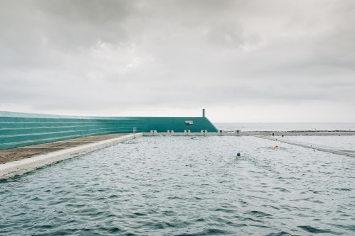 horizontal shot of a person swimming in a large ocean pool on a cloudy day