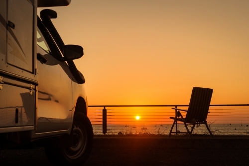 Horizontal shot of a white camper van at sunset in front of the sea with a foldable chair - Australian Stock Image