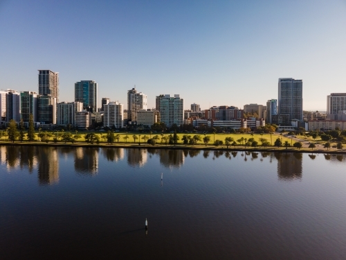 horizontal shot of buildings with trees and reflections appearing in a lake under clear blue skies - Australian Stock Image