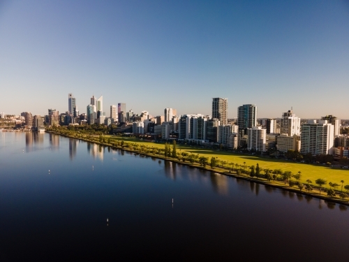 horizontal shot of buildings with trees reflecting in a lake under clear blue and white skies - Australian Stock Image