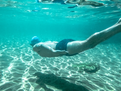 horizontal shot of man swimming underwater wearing trunks and swimming cap - Australian Stock Image