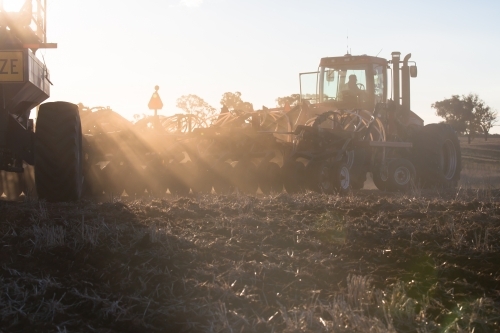 horizontal shot of silhouettes of tractors plowing soil in a sunny day