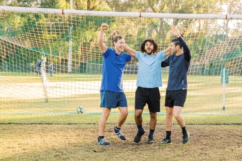 horizontal shot of three men smiling while raising their arms with goal in the background - Australian Stock Image