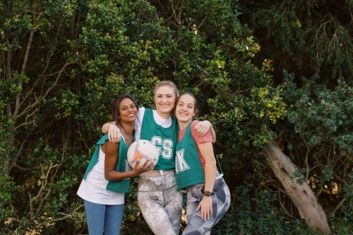 horizontal shot of three young smiling women posing outdoors with one holding a net ball - Australian Stock Image