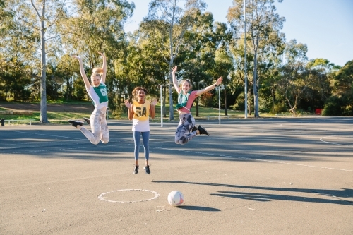 horizontal shot of three young women jumping in mid air with trees at the back and ball on ground - Australian Stock Image