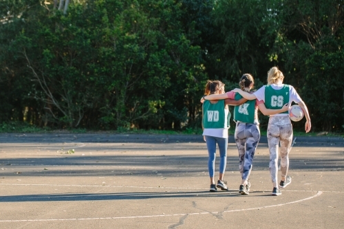 horizontal shot of three young women walking away with arms wrapped around each other on a sunny day - Australian Stock Image