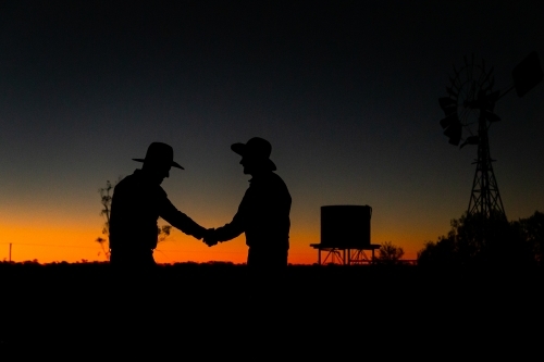 Horizontal shot of two men in silhouette wearing hats and shaking hands during sunset - Australian Stock Image
