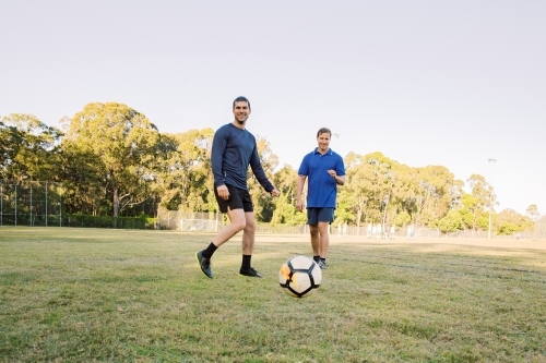 horizontal shot of two men playing soccer in a grass field with trees on a sunny day with clear sky - Australian Stock Image