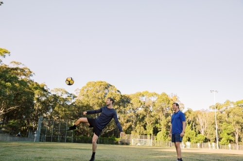 horizontal shot of two men playing soccer in the field with one man kicking the ball in mid air - Australian Stock Image