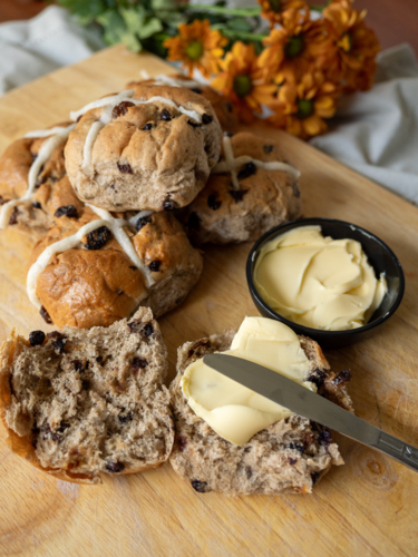 Hot cross buns on cutting board with butter and knife and orange flowers in background - Australian Stock Image