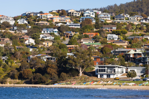 Houses densely scattered over a coastal hill with beach and water in the foreground at Blackmans Bay - Australian Stock Image
