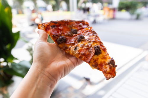 huge pizza slice held in a hand in an outdoor dining area - Australian Stock Image