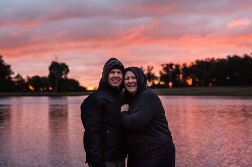 Husband and wife cuddling close together smiling happy with river water reflecting sunset sky