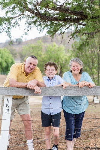 Husband and wife retired grandparents standing with grandson leaning on fence happy smiling