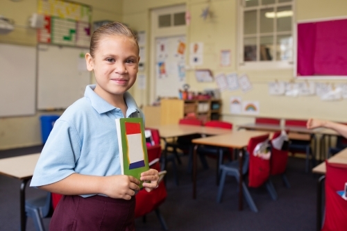 Indigenous primary school girl student holding books smiling in a classroom - Australian Stock Image