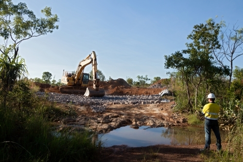 Industrial worker overseeing a large digger moving rocks on an industrial building site - Australian Stock Image