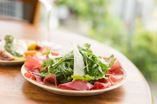 Italian bresaola, rocket and parmesan salad with a glass of wine and tomato salad in the background - Australian Stock Image