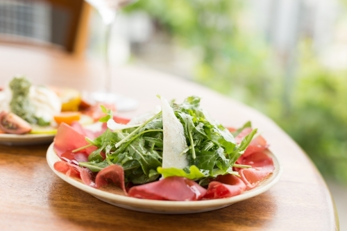 Italian bresaola, rocket and parmesan salad with a glass of wine and tomato salad in the background