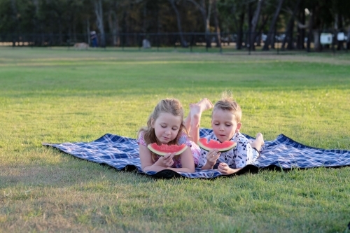 Kids eating watermelon on a picnic blanket in a park - Australian Stock Image