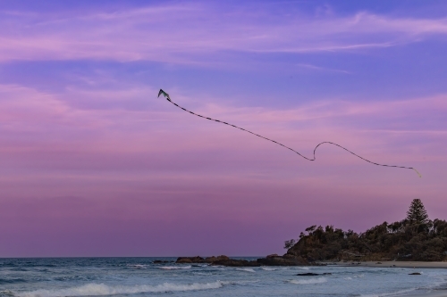 Kite flying over the beach through pink and purple sunset sky with long swirling tail - Australian Stock Image