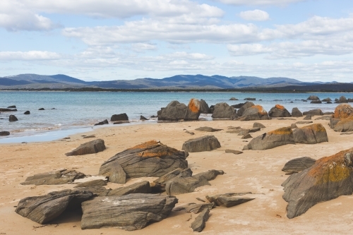 Large rocks along a beach, with mountain ranges and clouds in a blue sky