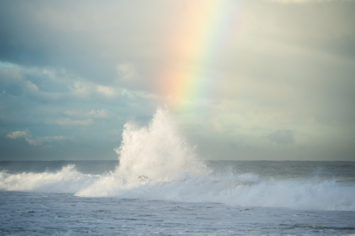 Large wave splashing into white spray with bright rainbow in background at Scarborough Beach, Perth - Australian Stock Image