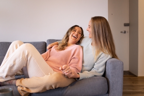 Laughing female same sex couple  in the living room, cuddling on the grey sofa
