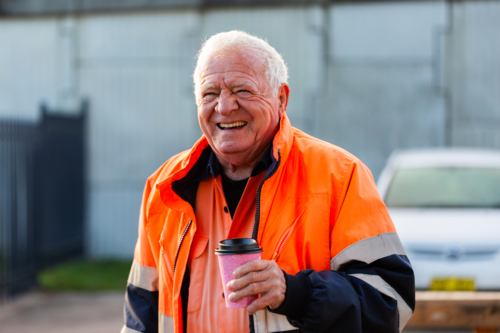 Laughing older Aussie workman in high vis on cold morning with warm takeaway coffee - Australian Stock Image
