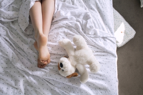 Legs and feet of a child laying in a messy bed with a teddy - Australian Stock Image