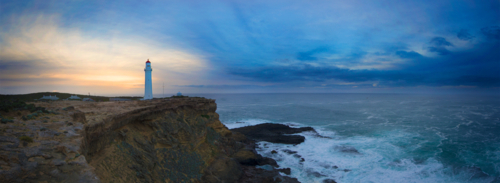 Lighthouse stands tall on a rugged coastal cliff at dusk. - Australian Stock Image