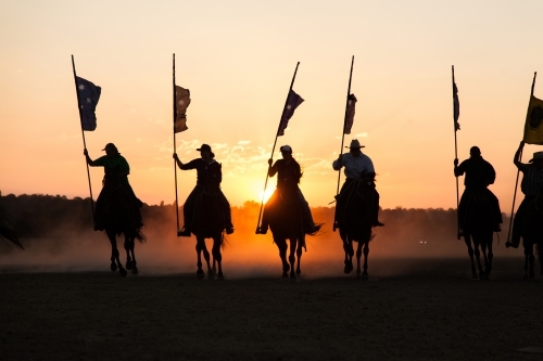 Line of silhouetted horses and riders with flag poles charging at sunset - Australian Stock Image