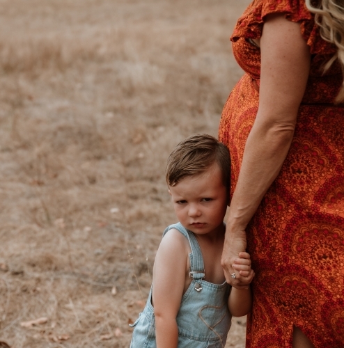 Little boy cautiously looking at the camera while leaning against his mother in a field