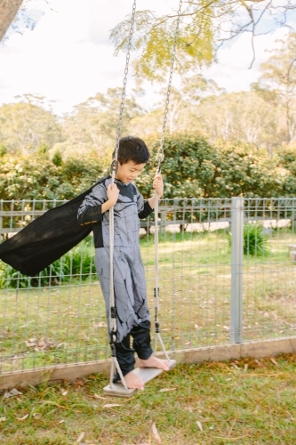Little boy dressed as batman playing in the garden on a swing on a beautiful day - Australian Stock Image