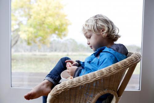 Little boy eating in wicker chair in front of window - Australian Stock Image