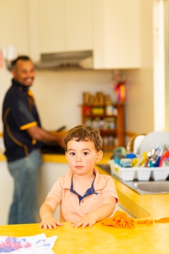 little boy standing at a counter playing with playdough with father in background in kitchen - Australian Stock Image
