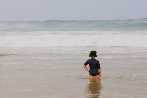 Little boy standing in shallow water at the beach watching waves roll in - Australian Stock Image