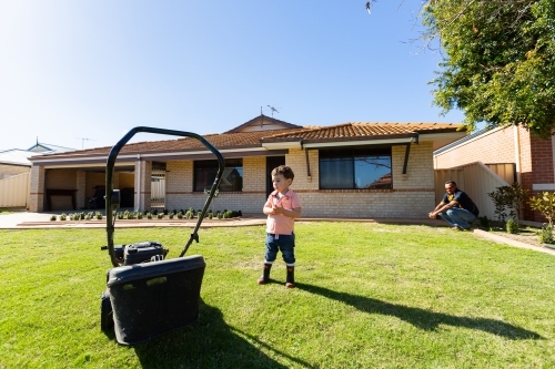 little boy standing on lawn near lawnmower watched by dad - Australian Stock Image