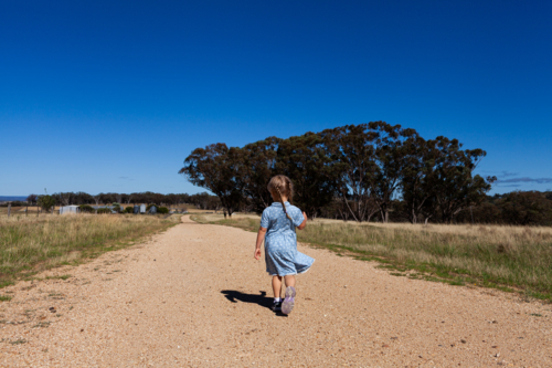 Little farm girl under blue summer sky running down gravel road through paddocks - Australian Stock Image