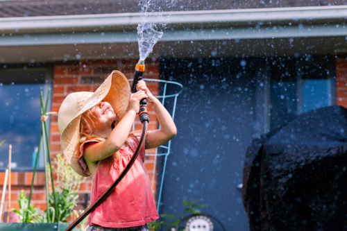 Little girl playing outdoors with hose in backyard wearing big floppy sun hat - water play - Australian Stock Image