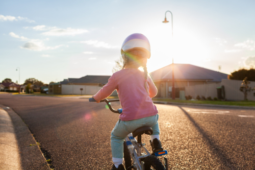 Little girl riding bike down quiet suburban road with backlighting at sunset - Australian Stock Image