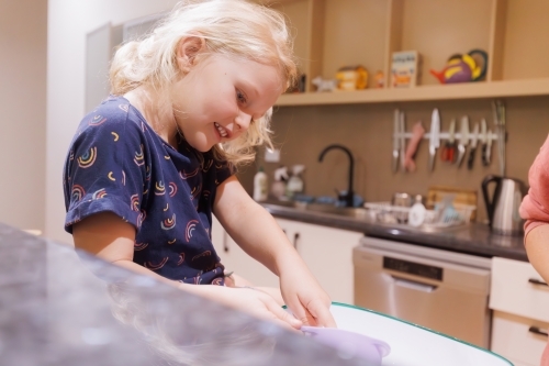 Little girl sitting on kitchen bench measuring ingredients into mixing bowl - Australian Stock Image