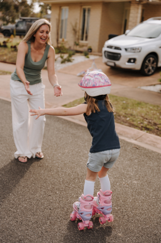 Little girl wearing pink helmet and roller skates reaching out to mum. - Australian Stock Image