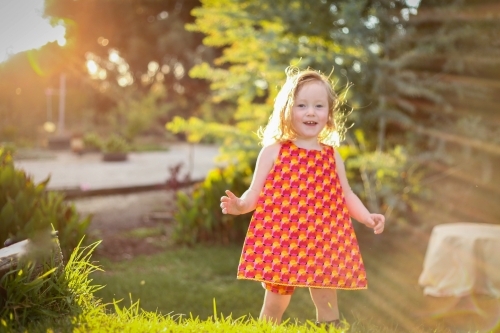 Little girl wearing red dress exploring garden flooded in golden afternoon light - Australian Stock Image