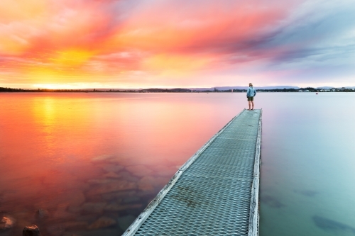 Lone figure standing on a jetty looking out at a vivid sunset - Australian Stock Image